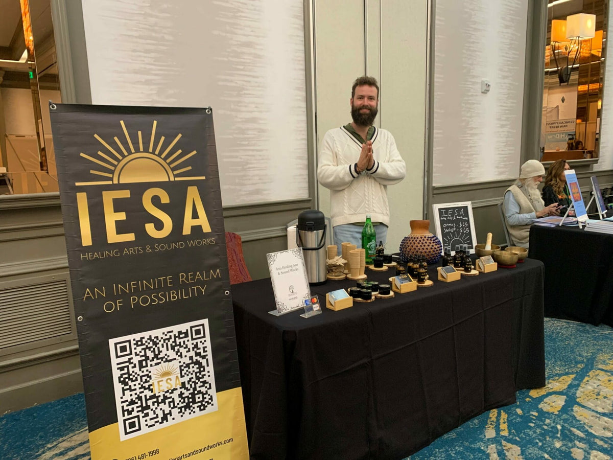 Clinton Greenlee standing behind the IESA Healing Arts & Sound Works table at an indoor event, with an IESA banner and products on display